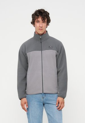 Jeune homme aux cheveux bouclés portant une veste polaire grise bicolore zippée et un jean bleu clair sur un fond blanc uni.