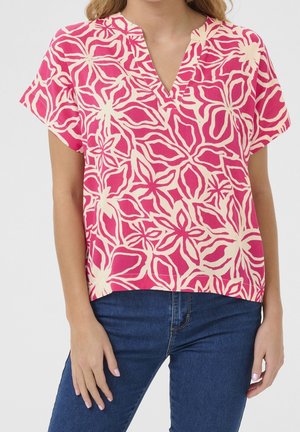 Kobieta w różowej bluzce z krótkim rękawem, kremowym kwiatowym wzorem i dekoltem w serek, w połączeniu z ciemnoniebieskimi dżinsami na białym tle.