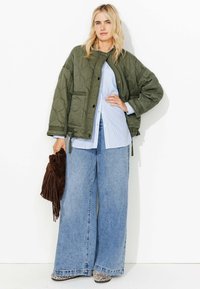 Veste matelassée verte avec fermeture à pression, chemise rayée bleu clair et jean large bleu. Tient un sac brun à franges et porte des chaussures à motifs.