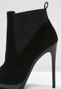 Stivale alla caviglia nero con tacco alto, realizzato in una combinazione di suede e materiale elastico, caratterizzato da un design elegante e una punta affusolata.