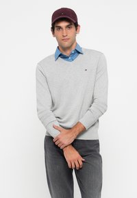Suéter cinza em V com punhos canelados, utilizado sobre uma camisa azul de colarinho. Boné burgundy. Modelo de pé com os braços cruzados, usando jeans cinza.