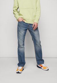 Sweat à capuche vert clair avec une poche avant, associé à un jean droit bleu délavé et des baskets multicolores avec des accents orange et blanc.