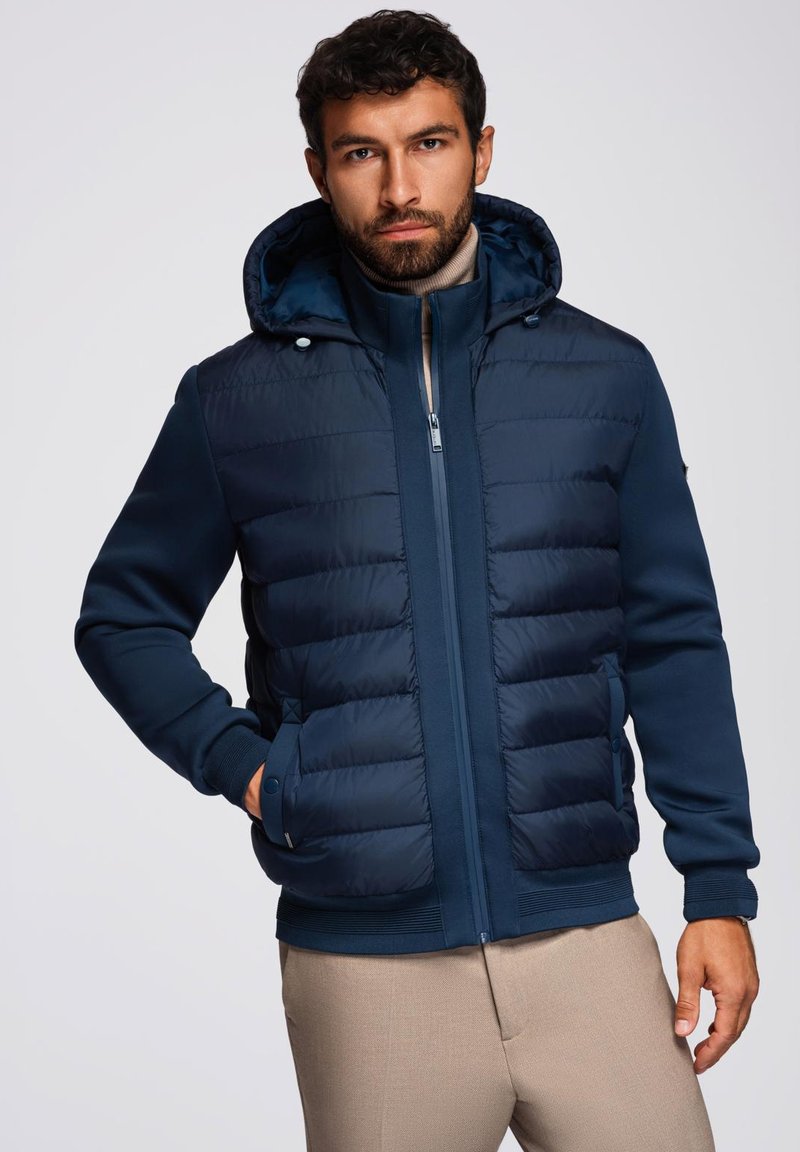 Giacca puffer blu navy con cappuccio, caratterizzata da sezioni trapuntate e maniche a tinta unita. Chiusura con zip, polsini a costine e tasche laterali.