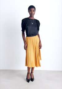 Haut en satin noir avec des manches bouffantes, associé à une jupe midi plissée jaune moutarde et des escarpins noirs à bout pointu. Collier simple en accent.