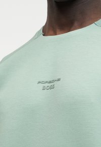 Κοντινό πλάνο ενός ατόμου που φοράει ανοιχτοπράσινο πουκάμισο με την επιγραφή "Porsche BOSS" εμφανιζόμενη σε τόνους πράσινου στο στήθος.