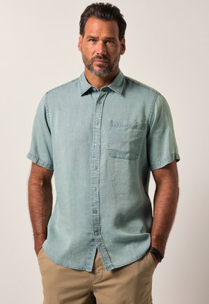 Mann mit Bart, der ein kurzärmliges hellblaues Hemd mit Knöpfen und beige Hosen trägt und mit den Händen in den Taschen vor einem schlichten Hintergrund steht.