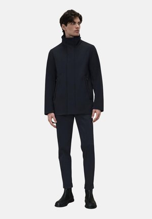 Giacca impermeabile blu navy con colletto alto, zip frontale e tasche laterali, abbinata a pantaloni neri slim e stivali neri alla caviglia.