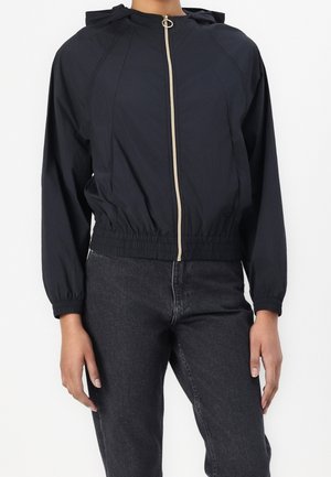 Veste noire à capuche avec fermeture éclair, ourlet et poignets élastiqués, portée avec un jean gris foncé.