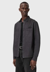 Uomo che indossa una camicia a bottoni grigio scuro sopra una maglietta nera e jeans neri, in piedi davanti a uno sfondo grigio chiaro uniforme.