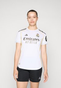 Maillot de sport blanc avec des accents noirs et jaunes, portant le texte "Emirates Fly Better", le logo du Real Madrid et une texture en mesh. Short noir.