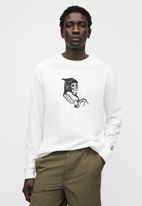 Λευκό μακρυμάνικο μπλουζάκι με μαύρο σχέδιο κρανίου και κείμενο «Volcom». Φορεμένο με χακί παντελόνι, με χαλαρή γραμμή.