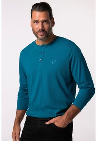 Tyrkysová dlouhá mikina s knoflíky typu henley, která má tři knoflíky u krku a decentní logo na hrudi, doplněná o černé kalhoty.