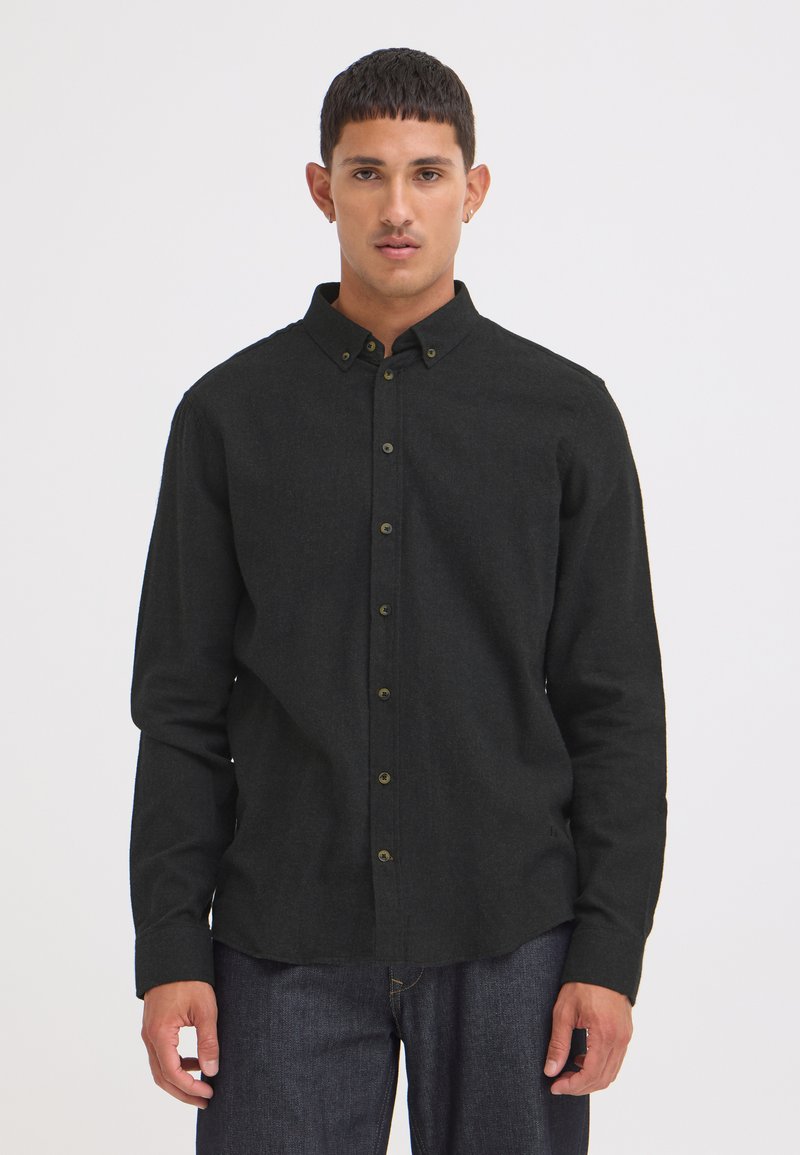Homme debout portant une chemise noire à manches longues boutonnée et un jean bleu foncé sur un fond clair uni.