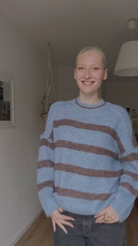 Mlada oseba se smeji, oblečena v modro jopico z rjavimi črtami, stoji v notranjosti ob steni z uokvirjeno umetnostjo in visečo rastlino.