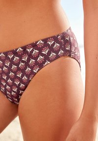 Bikiny s nízkym strihom v tmavej burgundskej látke s kvetinovým vzorom v bielej a ružovej farbe. Dizajn má hladkú textúru.