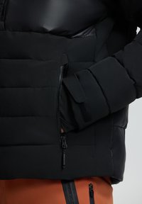 Chaqueta negra acolchada con textura acolchada, un zipper lateral y un bolsillo con solapa de Velcro en la manga. Acentos de tela suave y brillante.