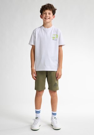 T-shirt blanche avec graphique vert, shorts olive, baskets blanches avec accents verts et chaussettes rayées bleues. Design simple, coupe décontractée.
