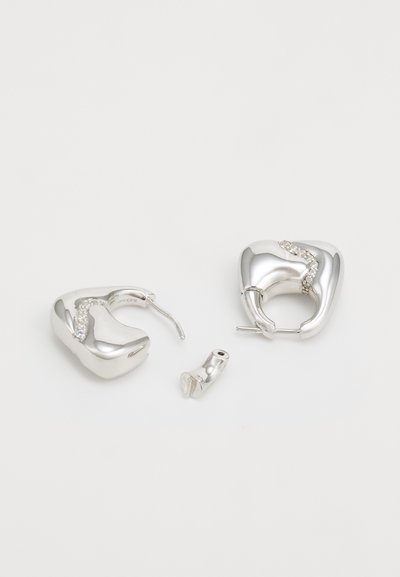 Silver hoop earrings with a smooth, rounded shape, featuring a clear crystal accent along a wavy design. One earring is detached.
