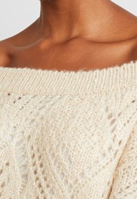 Sweter dzianinowy w kolorze kremowym, z dekoltem typu off-the-shoulder, charakteryzujący się luźnym krojem i skomplikowanymi wzorami koronkowymi, o miękkiej teksturze i prążkowanym dekolcie.