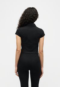 Γυναίκα με σγουρά σκούρα μαλλιά φοράει εφαρμοστό μαύρο πόλο με κοντά μανίκια και μαύρο παντελόνι, στέκεται γυρισμένη απέναντι σε λευκό φόντο.