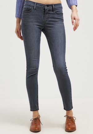 Femme portant un jean gris foncé ajusté, des chaussures marron à lacets et un pull court bleu, debout devant un fond clair uni.