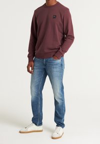 Bordowa bluza z okrągłym dekoltem i małym czarnym logo na piersi, w zestawie z jasnoniebieskimi dżinsami i białymi sneakersami z brązowymi akcentami.