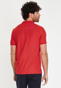 Camisa polo roja de manga corta, con un diseño sencillo y un acabado ligeramente texturizado. Presenta un cuello y un pequeño logo en el dobladillo.