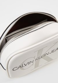 Borsa cosmetica in pelle bianca con cerniera argentata, caratterizzata da un logo "CK" in rilievo e un interno scuro a contrasto. Design compatto e rettangolare.