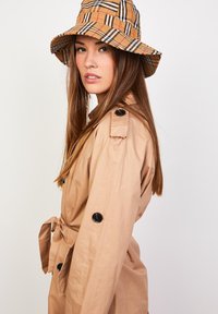 Abrigo trench beige con botones negros, cintura con cinturón y charreteras en los hombros, combinado con un sombrero tipo bucket a cuadros en tonos marrón y beige.