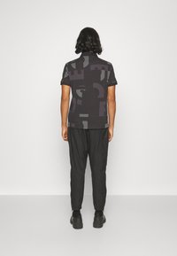 Černé polo triko s geometrickými vzory v tmavě šedé, spárované s černými zúženými kalhotami a mohutnými černými botami.