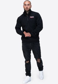 Felpa nera con zip e colletto alto, logo rosso sul petto, abbinata a jeans neri strappati e sneakers bianche.