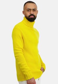 Leuchtend gelber Rollkragenpullover aus weichem Strick mit geripptem Saum und Bündchen, getragen zu weißen Hosen. Die Textur ist glatt und gemütlich.