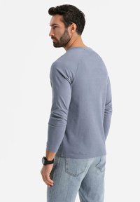 Långärmad blå ribbad t-shirt med rund halsringning, med en figurnära design och kontrasterande raglanärmar. Bärs med ljusblå jeans.