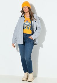 Chaqueta gris con cuello, camiseta gráfica amarilla, vaqueros azules y botines gruesos de ante beige. Un gorro de punto naranja completa el conjunto.