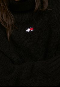 Person som bär en svart stickad tröja med ett litet Tommy Jeans-logotypmärke på bröstet. Långt brunt hår delvis synligt.