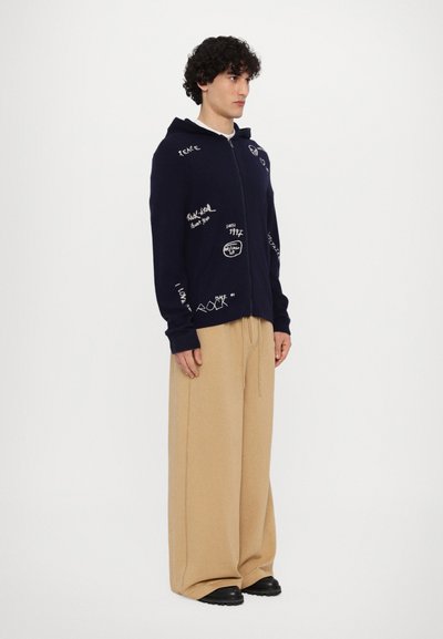 Haut à zip navy avec texte et graphiques manuscrits blancs, associé à un pantalon large beige et des chaussures noires. Texture douce.