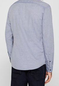 Camicia azzurro chiaro con tessitura a trama, maniche lunghe e orlo arcuato. Presenta bottoni scuri sui polsini.