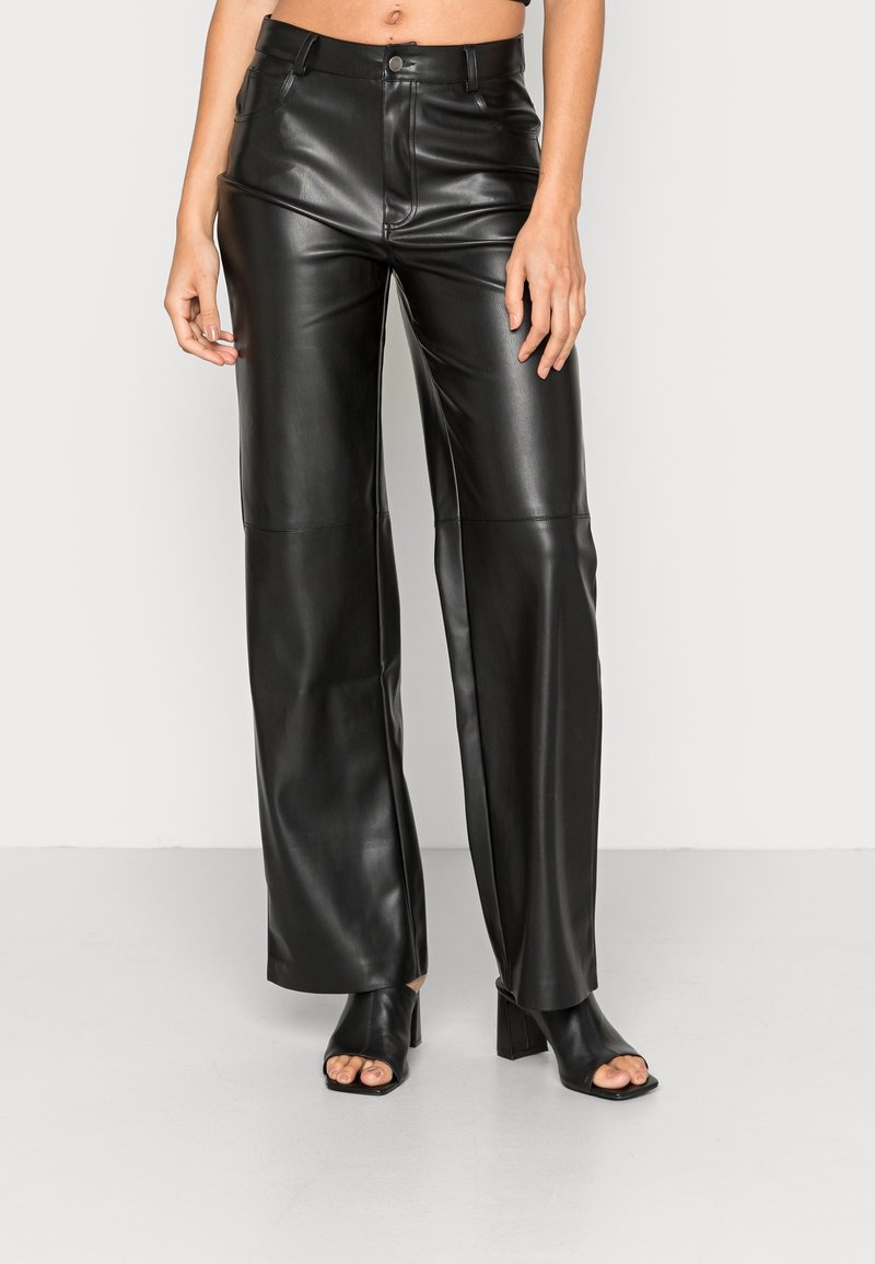 Femme portant un pantalon en cuir noir à taille haute et jambes larges, associé à des chaussures à talons blocs ouvertes noires, se tenant devant un fond neutre.