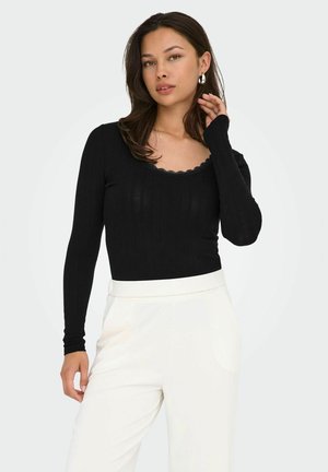 Schwarzes, geripptes Langarmoberteil mit einem gewellten Ausschnitt, kombiniert mit weißen High-Waist-Hosen. Glatte Textur, figurbetontes Design.