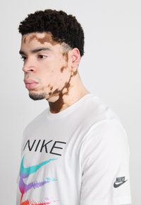 Balta Nike marškinėliai su juodu logotipu ir spalvingu grafiniu dizainu, kuriame yra mėlynos, purpurinės ir rožinės juostos. Modelis turi trumpus, garbanotus plaukus.