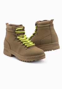 Botas de senderismo de cuero color marrón claro con una superficie texturada, cordones verdes, ojetes de metal y una suela de goma robusta con un dibujo en relieve.