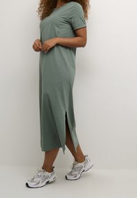 Jasnozielona sukienka midi z krótkimi rękawami i bocznymi rozporkami, wykonana z miękkiego materiału. W połączeniu z białymi i szarymi butami sportowymi.