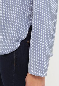 Gros plan d'une personne portant une chemise bleu clair avec des motifs de cœurs blancs et un jean bleu foncé, montrant la fente latérale de la chemise et les détails des poignets.