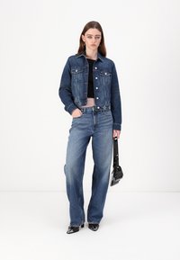 Džínová bunda v tmavomodrej farbe, s gombíkmi a vreckami. V kombinácii s svetlomodrými širokými nohavicami. Model drží malú čiernu tašku.