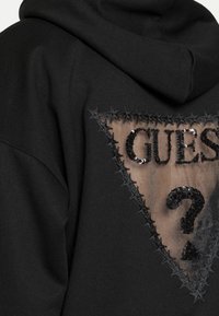 Sweat à capuche noir avec un panneau en maille transparente triangulaire orné du texte "GUESS?" en sequins et des coutures à motifs d'étoiles autour du bord.