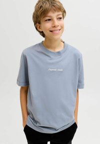 Νεαρό άτομο που χαμογελά, με κοντά μαλλιά, φορώντας ένα ανοιχτό μπλε μπλουζάκι "Originals Studio", με τα χέρια στις τσέπες, στέκεται μπροστά σε μια απλή φόντο.