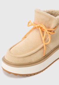 Botín de ante beige hasta el tobillo con cordones naranjas, interior acolchado, suela plataforma blanca gruesa y suela de goma texturizada marrón.