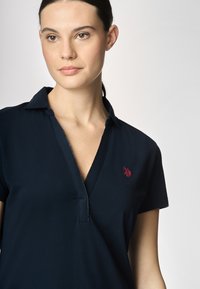 Polo shirt blu navy con colletto e maniche corte. Presenta un logo ricamato rosso sul lato sinistro del petto e una zip frontale. Texture liscia.