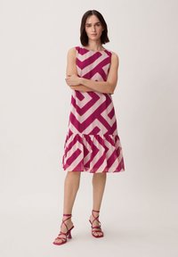 Ein rosa und cremefarbendes A-Linien-Kleid mit geometrischen Mustern, rüschigem Saum und ärmellosem Design. Kombiniert mit rosa Riemchen-Heels.