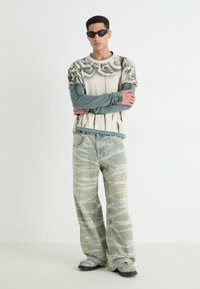 Overdimensjonert beige grafisk t-skjorte med svarte design lagd over en steinblå langermet topp. Lys blå mønstrede vide jeans. Sorte solbriller.